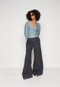 Blusa azzurro chiaro con maniche a sbuffo e fronte arricciato, abbinata a jeans larghi grigio scuro. Modella con occhiali da sole neri e tacchi.