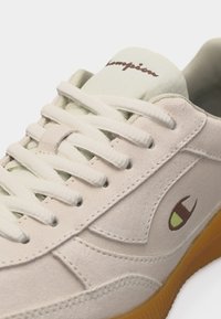 Μπεζ σουέντ αθλητικό παπούτσι με λευκά κορδόνια, καφέ και πράσινο λογότυπο "C" στο πλάι και σόλα από καουτσούκ χρώματος τσίχλας.