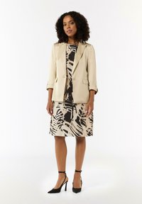 Beige maßgeschneiderte Jacke über einem schwarz-beige gemusterten Kleid mit kurzen Ärmeln und schwarzen spitzen Absatzschuhe mit Knöchelriemen.