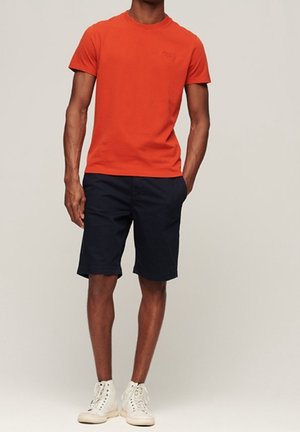 Man draagt een feloranje korte mouw T-shirt, donkerblauwe knielange shorts en witte hoge sneakers, staat met één hand in de zak.