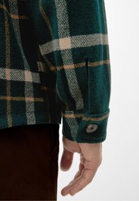 Camicia di flanella a quadri verdi con polsino abbottonato di un marrone accentuato. Il tessuto ha un aspetto morbido e texturizzato con linee beige.