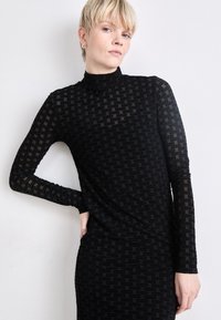 Black long-sleeve dress with a high neckline, made from sheer material featuring a repeating textured logo pattern. Form-fitting silhouette.