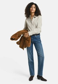 Skjorta med knappar i ljusbeige, bärande blå raka jeans och mörkbruna ankelboots, håller en jacka i brunt mocka.