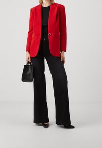 Blazer rouge sur mesure avec un seul bouton, associé à un jean noir à jambes larges. Tient un petit sac à main noir ; des chaussures noires pointues complètent le look.