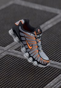 Nike running shoe featuring a gray mesh upper, black synthetic overlays, and orange accents. Rounded outsole with distinct cushioning design.