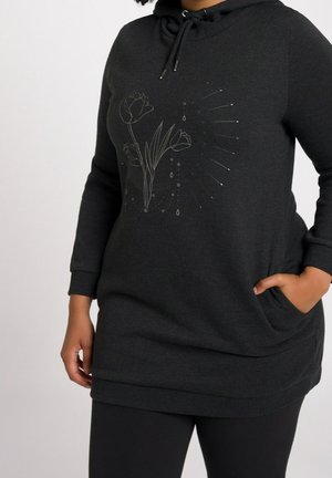 Personne portant un long sweat à capuche noir avec des cordons, un motif floral discret sur la poitrine et une main dans la poche.