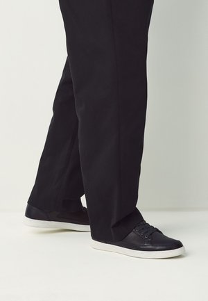 Personne portant un pantalon droit noir et des baskets noires avec des semelles blanches, debout sur un sol clair.