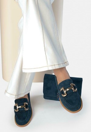 Mujer con pantalones blancos mostrando mocasines de ante azul marino con detalles dorados en forma de estribo, con bolso a juego de ante azul marino en el suelo.