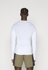 Långärmad vit träningsskjorta med meshtextur och slim fit-design. Kombinerad med olivgröna shorts med ett mörkt mönstrat sidopanel.