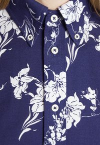 Marinblå skjorta med ett blommigt vitt tryck, med krage och knappar. Tyget har en strukturerad yta med en subtil lyster.