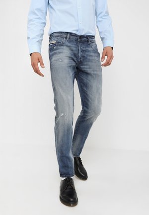 Man in vervaagde blauwe jeans met lichte slijtage, een lichtblauw overhemd met knopen en zwarte veterschoenen, staand tegen een witte achtergrond.
