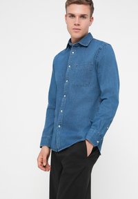 Džínsová košeľa v stredne modrej farbe, s gombíkovou prednou časťou, jednou náprsníkovou vreckom a dlhými rukávmi. Hladká textúra s klasickým golierom.