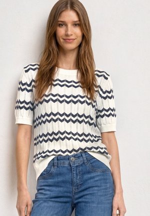 Jeune femme aux longs cheveux bruns portant un pull en maille blanc avec des rayures zigzag noires et un jean bleu, debout contre un mur blanc.