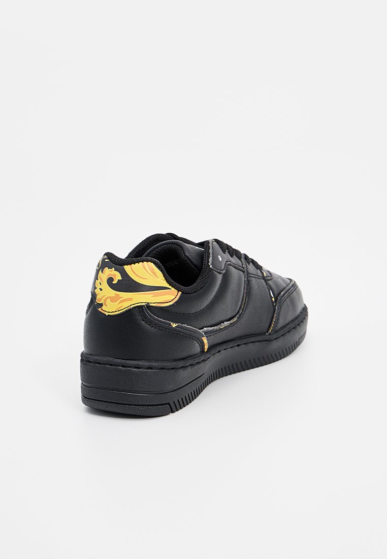 Zapatilla de cuero negra con una superficie texturizada, que presenta un audaz diseño ornamentado amarillo en el talón y una suela de goma duradera.
