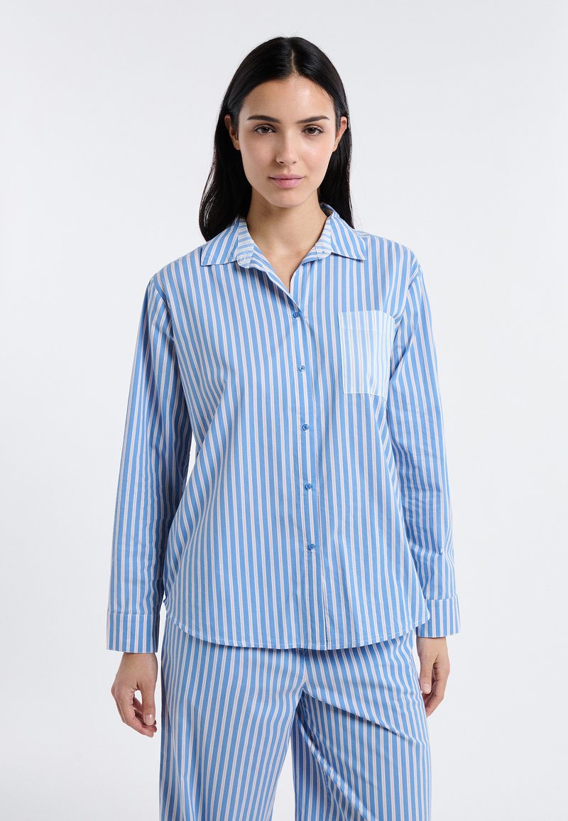 Femme portant une chemise à manches longues et un pantalon assortis à rayures verticales bleues et blanches, debout devant un fond blanc uni.