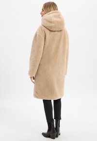 Frau in einem beigefarbenen, flauschigen Kapuzenmantel, schwarzen Hosen und schwarzen Absatzstiefeln, die seitlich nach links vor einem weißen Hintergrund steht.