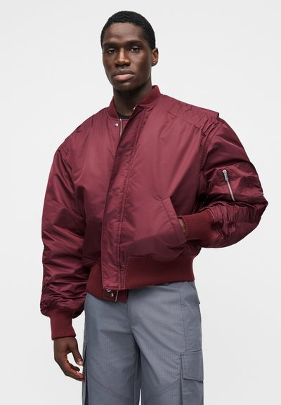 Bordová bomber bunda s rebrovanými manžetami a golierom, vybavená vreckom na zips na rukáve a hladkou, lesklou textúrou.