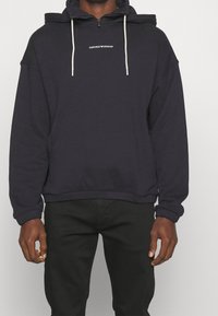 Mörkblå zip-up hoodie med fleece-textur, utrustad med dragsko på huvan och vit logotyp på bröstet. Matchas med svarta byxor.