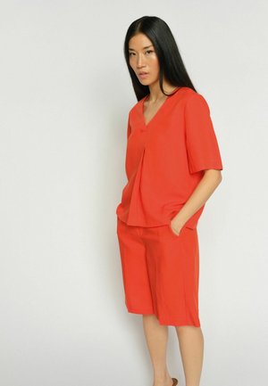 Pikkade mustade juustega naine, kes kannab ereoranži V-kaelusega lühikese varrukaga topi ja sobivaid põlvekõrguseid lühikesi pükse, seisab ühe käega taskus.