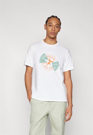 Camiseta de algodón blanca con un gráfico circular en verde y naranja que presenta el texto "Converse Chuck Taylor All Star." Mangas cortas, escote redondo.