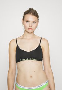 Bralette nera con spalline sottili, con logo Calvin Klein verde sulla fascia. Tessuto liscio con design a scollo a V.