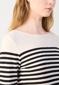 Stripet langermet topp som har en kremfarget øverste del og svarte striper på den nedre halvdelen. Stoffet har en glatt tekstur og en båtnek. 