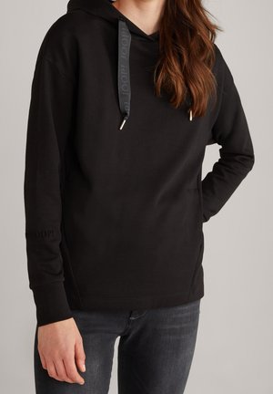 Femme portant un sweat à capuche noir avec cordons et un jean foncé, debout avec une main sur la hanche devant un fond uni.