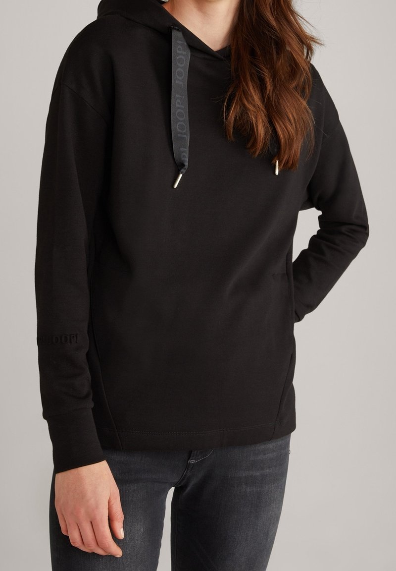 Femme portant un sweat à capuche noir avec cordons et un jean foncé, debout avec une main sur la hanche devant un fond uni.