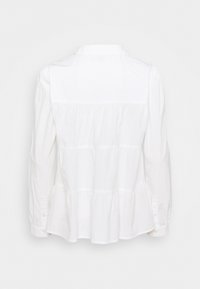 Blouse blanche à manches longues avec détails à volants superposés dans le dos et poignets boutonnés, présentée sur un fond uni.