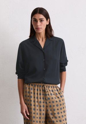 Donkergrijs overhemd met kraag, met opgerolde mouwen. Gekozen in combinatie met een lichtbruine broek met trekkoord en zwarte accenten.