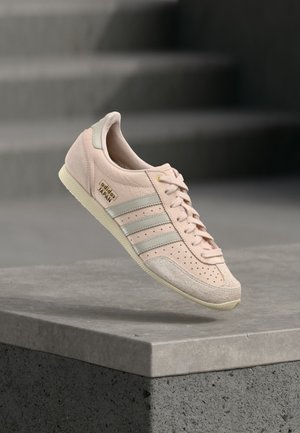 Ροζ ανοιχτό αθλητικό παπούτσι Adidas Japan με γκρι ρίγες και μπεζ σόλα, να αιωρείται πάνω από μια γκρι τσιμεντένια άκρη με σκάλες στο φόντο.