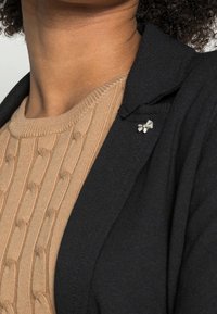 Czarna marynarka na teksturowanym beżowym swetrze, z małą srebrną kokardką przypiętą do klapy marynarki.