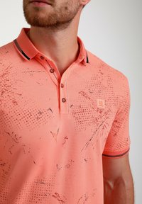 Koralo spalvos polo marškiniai su tekstūruotu raštu, dekoratyviomis apykaklės juostelėmis ir logotipo aplikacija. Turi klasikinę apykaklę ir dviejų mygtukų užsegimą.