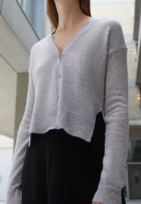 Helle graue, kurz geschnittene Strickjacke aus weichem Material mit V-Ausschnitt, vorderer Knopfleiste und gerippten Säumen, kombiniert mit schwarzen Unterteilen.