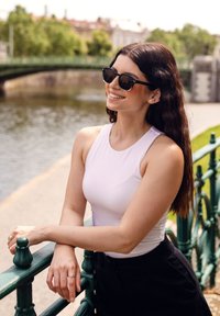 Bílý bezramenný top, černé sluneční brýle a černé kalhoty. Stojí u zeleného zábradlí, v pozadí voda a zeleň.