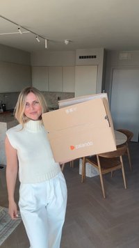 Zalando kartonska kutija, svijetlosmeđa s narančastim logotipom, drži osoba. Unutrašnjost prikazuje modernu kuhinju sa svijetlim drvenim podom i namještajem.