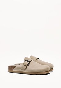 Pár béžových semišových slip-on drevákov s nastaviteľnými čiernymi prackami a plochou korkovou podrážkou na bielom pozadí.