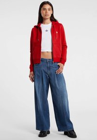 Giovane donna che indossa una felpa rossa con zip, una t-shirt bianca corta con stampa di stelle, jeans a gamba larga blu e scarpe nere, in piedi davanti a uno sfondo semplice.