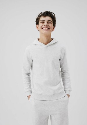 Lichtgrijze hoodie van zacht materiaal, met een buidelzak, dunne koordjes en ribbelmanchetten. Model staat met handen in de zakken.