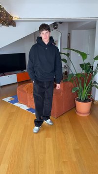 Černá mikina s kapucí, černé široké kalhoty a stříbrné tenisky. Umístěno uvnitř, na pozadí květináč s rostlinou a texturovaná oranžová pohovka.