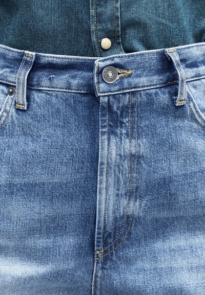 Κοντινό πλάνο της μέσης τζιν παντελονιού σε μπλε τζιν ύφασμα με κουμπί και θηλιές ζώνης, φορεμένο πάνω από ένα πιο σκούρο μπλε τζιν πουκάμισο.
