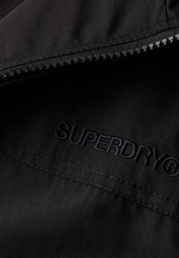Sort tekstil med en glat overflade prydes af et broderet "SUPERDRY" logo. Lynlås lukningen har et robust sort lynlåsdetalje.