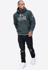 Sudadera verde oscuro con un bolsillo tipo canguro y un gran logo en la parte frontal. Combinada con jeans negros desgastados y zapatillas blancas de diseño alto.