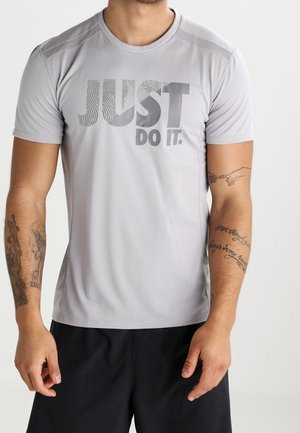 Mężczyzna w jasnoszarej koszulce z napisem "JUST DO IT." oraz czarnych szortach, prezentujący tatuaże na obu przedramionach na jednolitym tle.