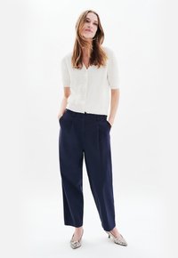 Vit kortärmad cardigan med broderade detaljer, matchad med marinblå vida byxor och mönstrade klackar, stående mot en ljus bakgrund.