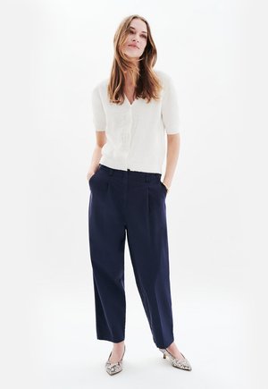 Weißer kurzärmeliger Cardigan mit besticktenDetails, kombiniert mit marineblauen weiten Hose und gemusterten Absatzschuhen, vor einem hellen Hintergrund stehend.