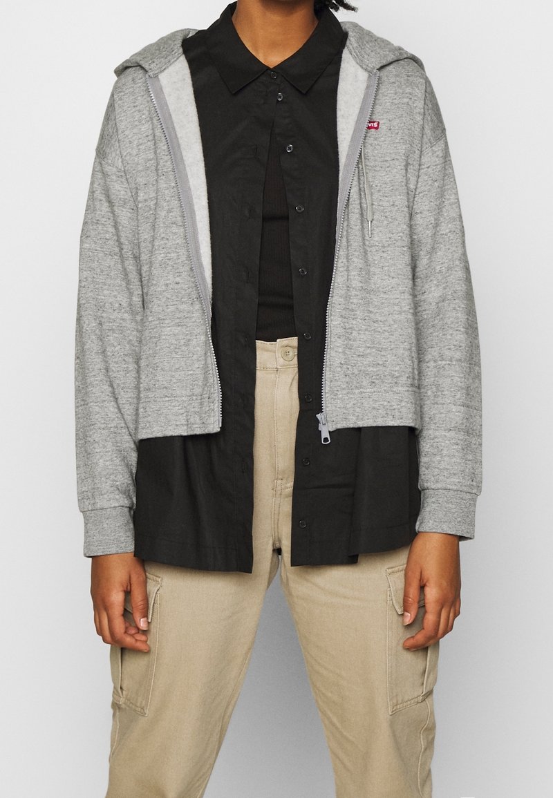 Personne portant un pantalon cargo beige, une chemise noire boutonnée sur un haut noir, et un sweat à capuche gris zippé avec une petite étiquette rouge sur la poitrine.