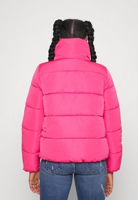 Person med två flätade svansar som bär en ljusrosa pufferjacka och blå jeans, visad bakifrån mot en neutral bakgrund.