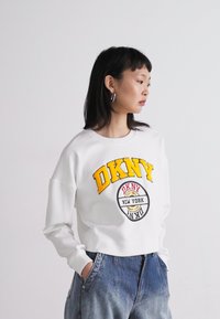 Λευκό κοντό φούτερ με έντονα κίτρινα και μαύρα γράμματα του λογότυπου "DKNY" και κυκλικό διακοσμητικό "NEW YORK"; φοριέται με ανοιχτό μπλε τζιν παντελόνι.