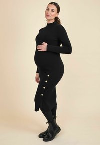 Abito maternità a costine nero con collo alto, maniche lunghe, spacco laterale e dettagli in oro. Indossato con stivaletti neri alla caviglia.